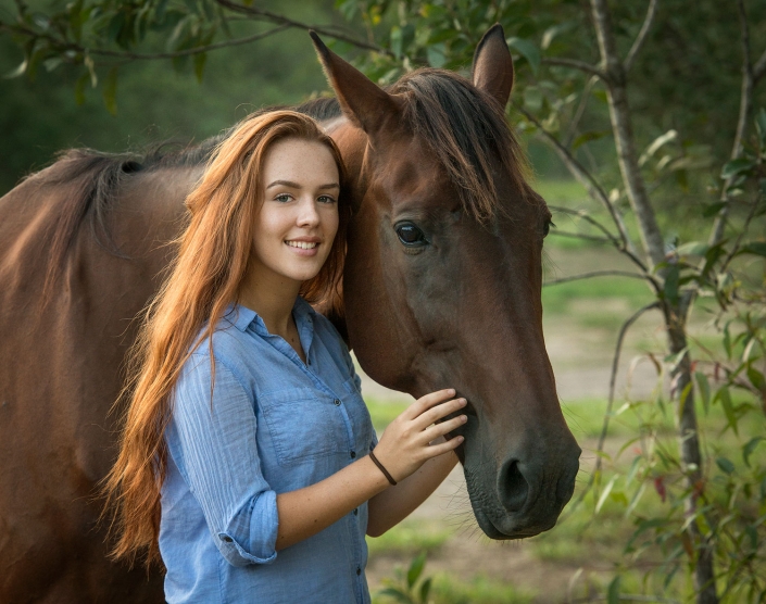 beautiful red hair girl standing with brown horse smiling