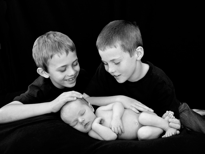 black and white photograph of two young brothers smiling at newborn baby sibling wearing black tshirt
