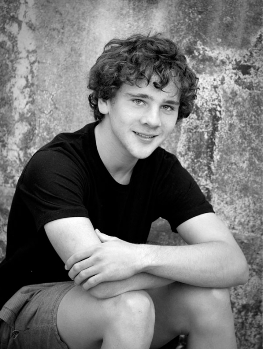 black and white image of curly haired teenage boy wearing black t shirt and folding his arms