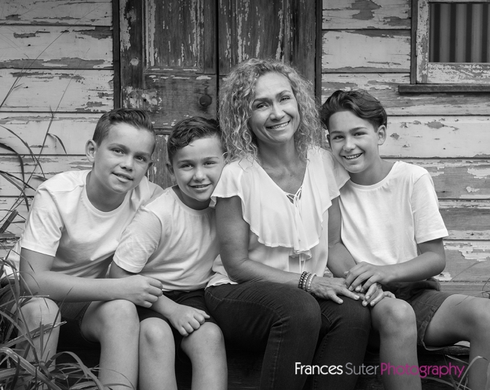 Mother and her three sons pose in front seated in front of rustic wood panel shed wearing white t-shirts, jeans and short