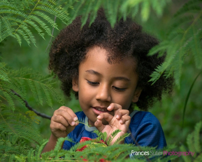 candid image of curly haired boy playing with leaf in garden fern