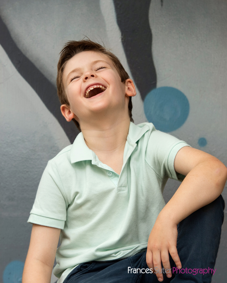 happy young boy wearing teal coloured t shirt laughs while posing for portrait in front of street art background
