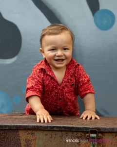 Toddler boy leans on large metal box laughing wearing red shirt