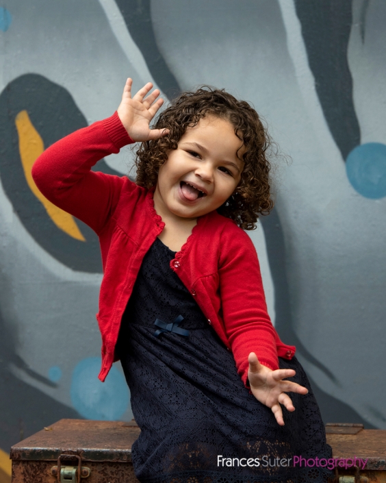 Young girl with curly hair laughing and striking a funny pose with hands in air wearing red cardigan and navy blue dress in front of graffiti wall