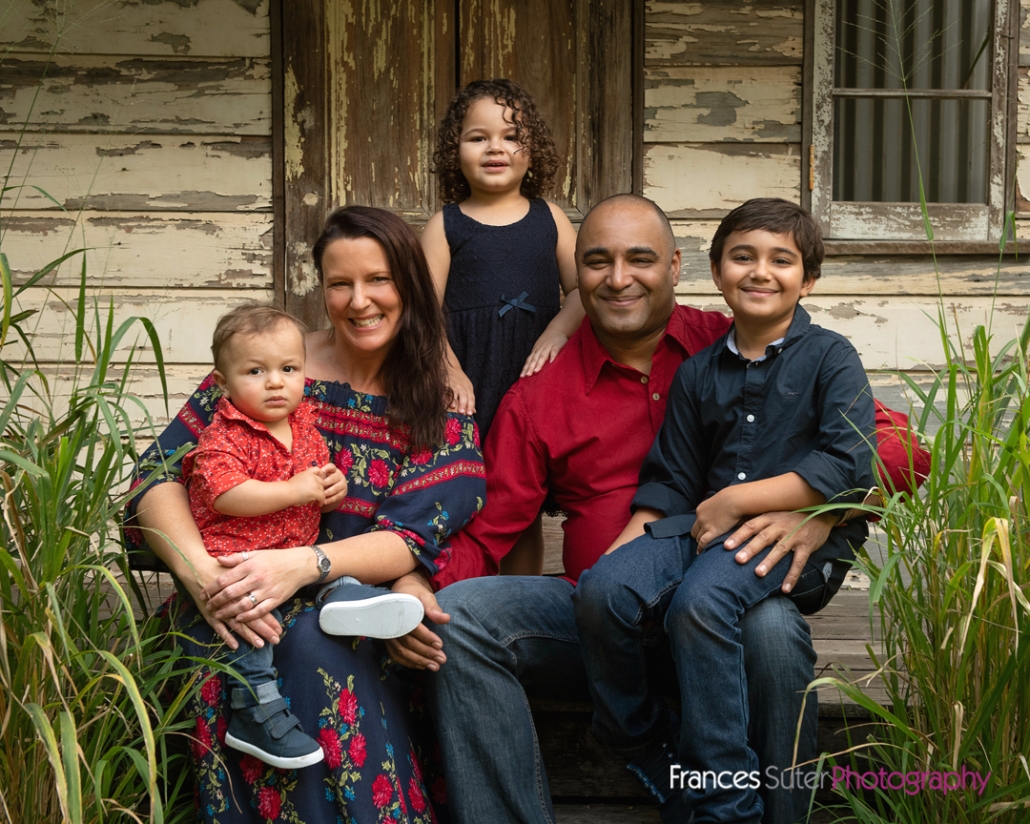Family of five, Mother, father, two sons and daughter wearing red and navy blue sitting in front of rustic shed backdrop
